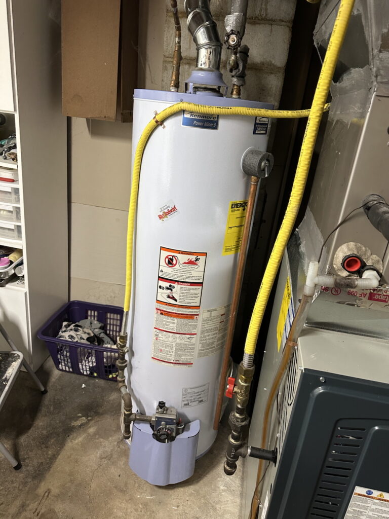 An upright, cylindrical Kenmore Power Miser 9 gas water heater painted white, featuring a yellow EnergyGuide label and warning stickers on its front, connected to copper and flexible yellow gas lines.